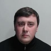 White man in 30s with round face and short dark hair wearing black zip-up jumper