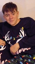 An image of a white teenage boy with short light brown hair, wearing a black top with white antlers and multi-coloured christmas lights on it, leaning in front of a white wall