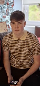 Teenage boy sitting on a chair in front of a decorated Christmas tree. He has short dark hair combed to one side. He is wearing a white collared, checked top and dark coloured trousers. He is holding a mobile phone in his hand.