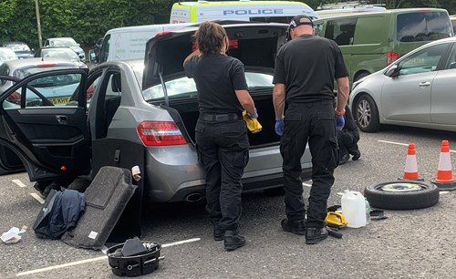 Image shows a busy car park with a police van in the background. In the foreground, two officers are searching the boot of a silver car. The doors of the car are open and there are various items on the ground, including a spare tyre and two orange cones.