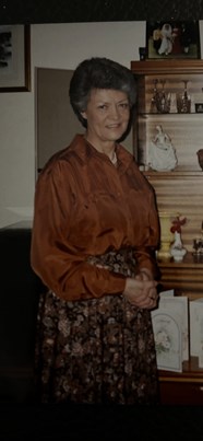 Woman who has short hair and is wearing a rust-coloured shirt, pearls and a floral skirt. She is standing in front of a dresser unit with her handd clasped in front of her.