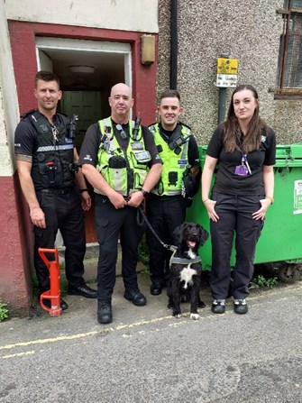Image of officers, trading standards dog handler and dog, and trading standards officer standing outside doorway in street. Officer on the left wearing black, dog handler and another officer wearing hi-vis vests, and female trading standards offier on right wearing all black.