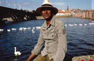 Man wearing white hat and shirt sitting on a boat with water and swans behind him.