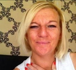 Woman with short blond hair. She is looking straight ahead and sitting on a dark coloured chair. Behind her is black and white patterned wallpaper. She has her hand at her neck and is wearing red nail polish.