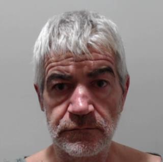 Image shows a man. He has shaggy grey hair, a short grey beard. He has deep lines on his forehead.