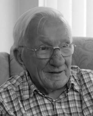Black and white photograph of an elderly man with short white hair, smiling, wearing glasses and a checked shirt sitting on a couch