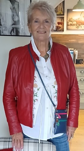 68-year-old woman with short hair smiling for the camera. Wearing a white shirt, red jacket and jeans.