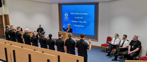 Police Scotland probationary constables in black uniforms standing with backs to camera making their oath of office at Tulliallan, watched by senior officers in formal uniforms with white shirts and black trousers.