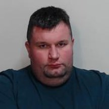 Image of Leigh Sutherland - white man, heavy build, with short dark hair and dark stubble wearing a navy blue t-shirt