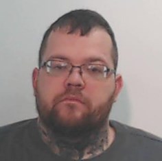 Head and shoulders photograph of a man in his 30s with shirt dark hair, thick beard and glasses. He's wearing a t-shirt and looking at the camera