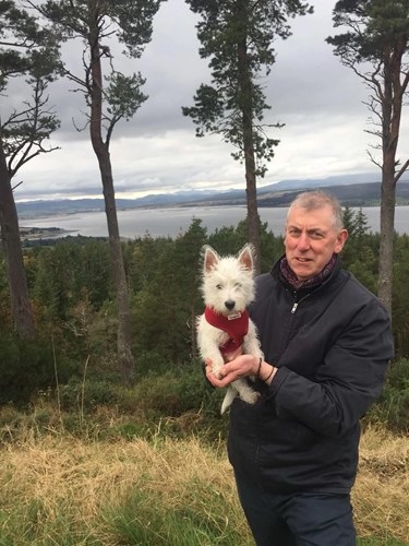Picture of David MacKenzie - older white man with short grey hair wearing dark coloured jacket, trousers and scarf holding a west highland terrier puppy in front of trees, a body of water and mountains in the distance.