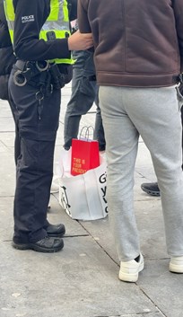 Image of officer holding man wearing brown leather jacket and grey jogging bottoms by the elbow on street with bag of shopping on the ground