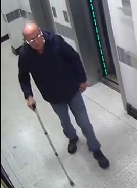 Bald white man walking in hallway with walking stick wearing a dark-coloured jacket, trousers and shoes. There is an elevator to the right of the picture.