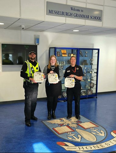 Police officer with school support staff member and SFRS colleague holding training defibrillators in front of Musselburgh Grammar School sign