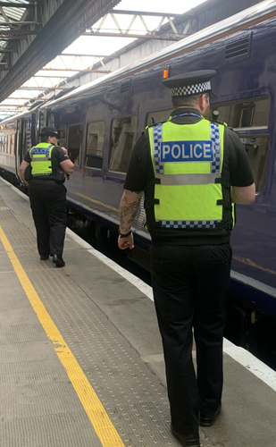Two uniformed officers are walking along the platform of a train station and there is a dark blue train to the right.