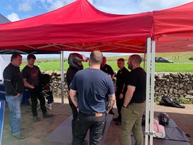 Image of seven men standing under a red gazebo learning about motorcycle safety with one man in the centre wearing a helmet and protective clothing.