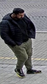 Man with black hair and beard. He is wearing a black top and jacket and trousers