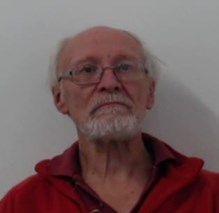 Man wearing glasses with grey hair at the sides and bald in the middle is looking straight ahead. He has a grey moustache and beard and is wearing a red top