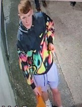 White male with brown hair. He is wearing a Nike jacket which is black at the top and multi-coloured from the chest down, including green, pink and yellow colours. He is wearing lilac coloured shorts. He is pictured in the street.