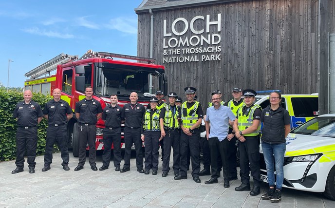 Officers from Scottish Fire and Rescue Service, Police Scotland, and British Transport Police with Councillor David McBride of West Dunbartonshire Council and Ranger Service Manager Leigh Hamilton from Loch Lomond and The Trossachs National Park, standing in front of a fire engine, police cars and the Loch Lomond & The Trossachs National Park building.