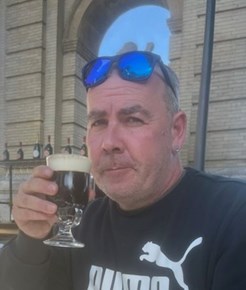 Man wearing a black long sleeved top with sunglasses on his head holding a drink sitting outside