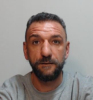 Image shows a man. He has short dark grey hair and beard. He has tanned skin. He is wearing a grey jumper and looking directly at the camera.