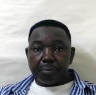 Custody image of Walter Masocha, black man with short hair and facial hair wearing white t-shirt and blue and white checkered shirt standing in front of white background