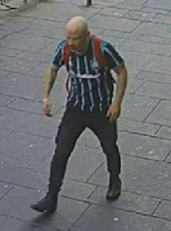 Image shows a white, bald man walking along a pavement. He is wearing a black and green striped football top, black jeans and black shoes. He is carrying a rucksack with red straps, which are visible.