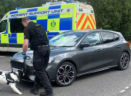 A dark silver car is being searched by an officer and a black and white dog. There is a police 'incident support vehicle' in the background.