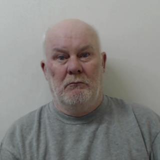 Image shows an older man who is balding and with white hair round the sides. He has a short, white beard and moustache. He is wearing a grey jumper.