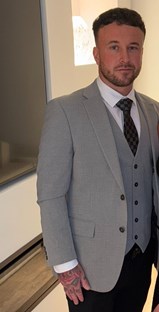 Man standing in front of a light with a white wall behind him. He has short dark hair, and short facial hair. He is wearing a light grey suit and dark coloured tie.