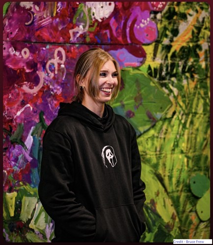 Image shows a young woman, smiling broadly. She is not looking off to the right. She is wearing a black hoodie with a white logo on the front. In the background is colourful graffiti.