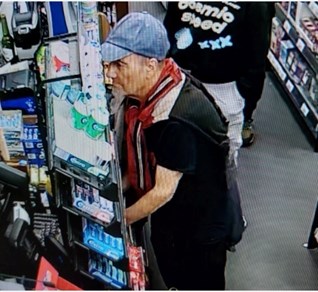Man within a shop at the till. He is wearing a light blue baseball cap with a red, black and white scarf. He appears to be paying for something. In the background there is a person standing behind him with a black sweatshirt and writing on the front. In front of him are rows of confectionary.