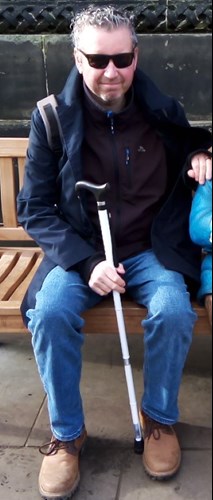 He is described as 6ft 4in tall, of slim build, with short grey hair. In the picture he is sitting on a bench wearing sunglasses, a black jacket, blue jeans, brown shoes and holding a white walking stick.