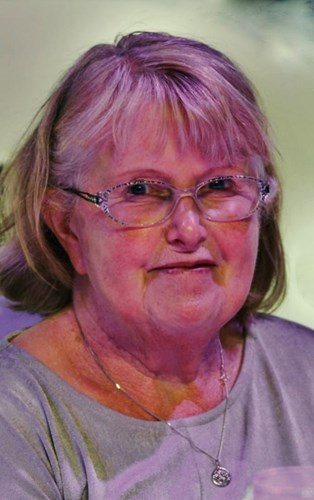 Image shows an older woman. She is wearing glasses and smiling slightly. Her hair is medium length and with a fringe.