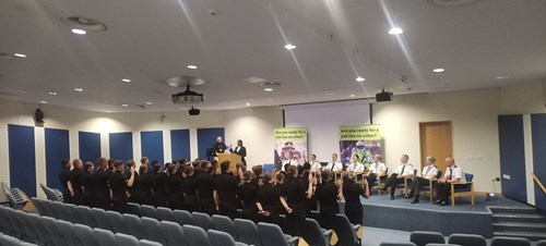 Police Scotland probationary constables in black uniforms standing with backs to camera making their oath of office at Jackton training centre, watched by senior officers in formal uniforms with white shirts and black trousers.
