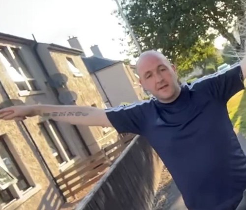 William Devaney, a white male standing in a garden in front of houses. He is bald and wearing a blue Adidas t-shirt