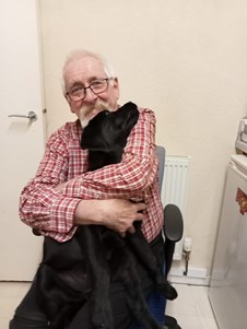 An image of a white man with white hair and beard, wearing dark coloured rimmed glasses and a red and white checked shirt. He is sitting on a grey computer chair, cuddling a black, labrador-type dog, in front of a cream coloured wall, white door and white radiator.
