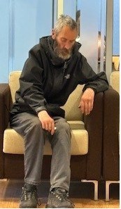Image of white man with grey hair and facial hair. Wearing a black jacket, grey trousers and black boots