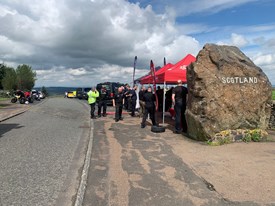 Image of police and fire service standing beside road talking to motorcyclists under red and blue gazebos. Rock with Scotland written on it in the foreground and vehicles and motorcycles in the background.