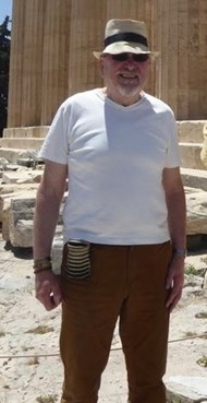 Man wearing a straw, sun  hat with a black band around it. He has a grey beard and is wearing sunglasses and a white t-shire. He is wearing brown trousers and is standing in front of marble pillars.