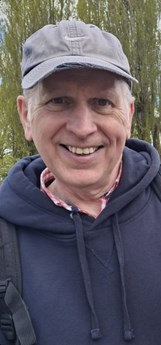 Man in his 60s wearing a grey baseball cap. He is standing in front of trees and smiling. He is wearing a blue hooded top and a red checked shirt.