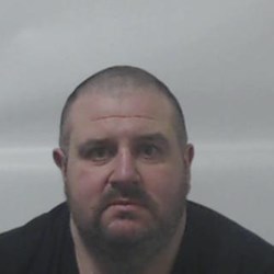 Man with very short dark hair and a beard. Wearing a dark coloured top.