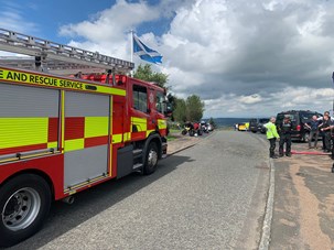 Image of fire engine beside a Scotland flag with police, fire service and motorcyclists in the background speaking. Further behind are vehicles and motorcycles parked at the side of the road.