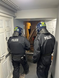 Two police officers wearing blue helmets and black clothing with the word Police written across their back. They are within a corridor in front of a property as part of an operation