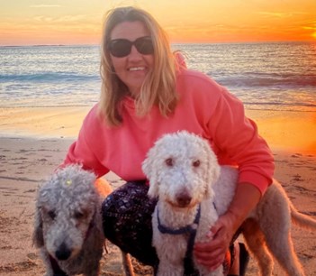 Women with blonde hair and red hooded jumper is kneeling on beach with her two dogs at sunset