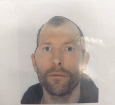 White man staring straight ahead. He is not smiling in the picture and has a short beard. He is wearing a dark coloured top.