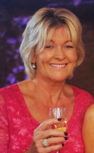 Image shows a woman with short, blonde hair. She is smiling broadly. She is wearing a pink, short-sleeved top.